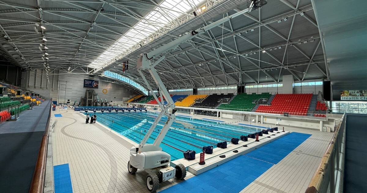 A large indoor swimming pool facility with tiered seating in multiple colours and a mobile boom lift positioned beside the pool.