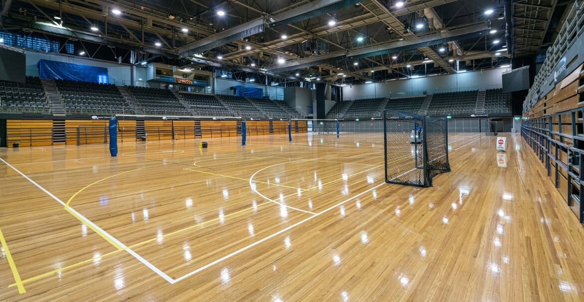 A large indoor sports arena with polished timber courts, multiple nets, and tiered spectator seating under a high industrial ceiling.