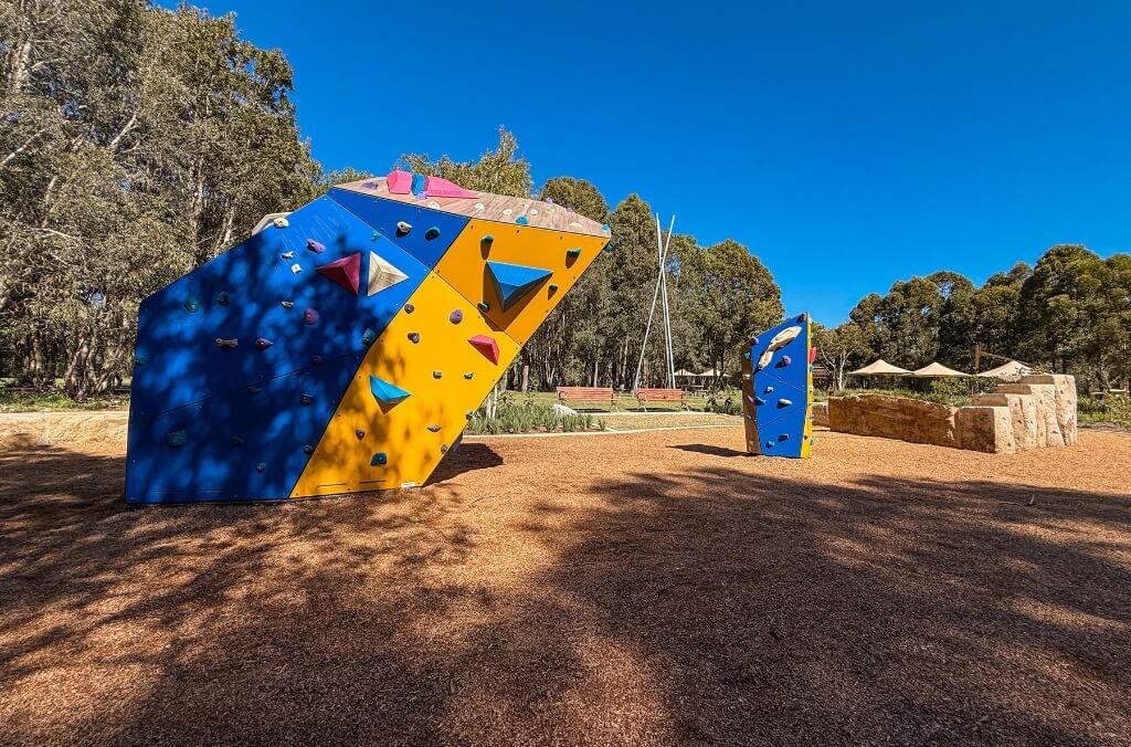 Outdoor climbing area featuring two artificial blue and yellow climbing structures with colorful holds, set against a backdrop of trees and a clear blue sky.
