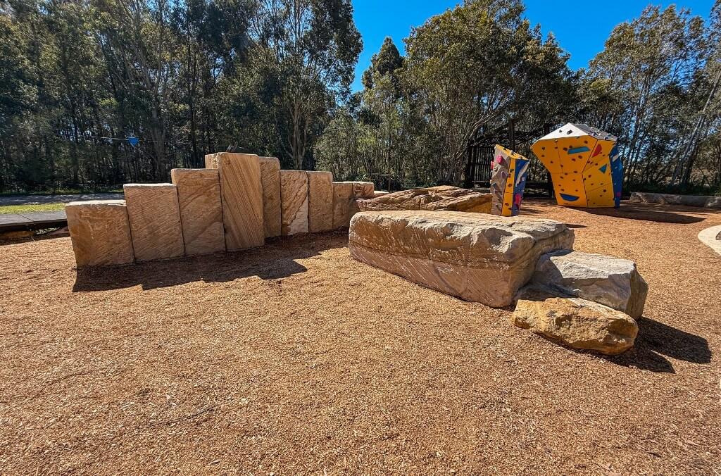 Outdoor area with large stone blocks arranged in a row, a flat stone in the foreground, and a colorful climbing structure with handholds in the background, surrounded by trees and mulch-covered ground.